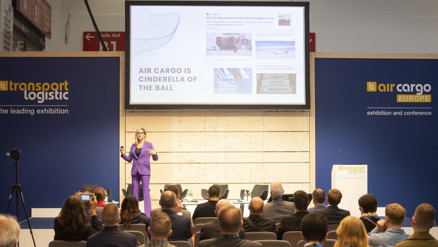 A woman in a purple suit gives a lecture to an audience. Behind her is a large screen and dark blue walls with the logos of 