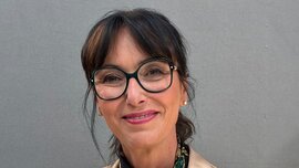A smiling woman with long dark hair and large black glasses is wearing earrings, a shimmering beige top, and a scarf adorned with jewelry.
