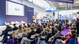 A lecture scene at a trade show: A speaker is presenting on stage in the “Forum Laboratory & Analysis” in front of a large audience.