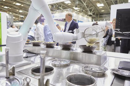 A robotic arm prepares food at a trade fair stand. There are filled bowls on a counter in front of it.