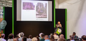 Publikum blickt auf eine Bühne auf der INTERFORST, auf der ein Mann mit Mikrofon eine Präsentationsfolie auf der Leinwand erläutert.