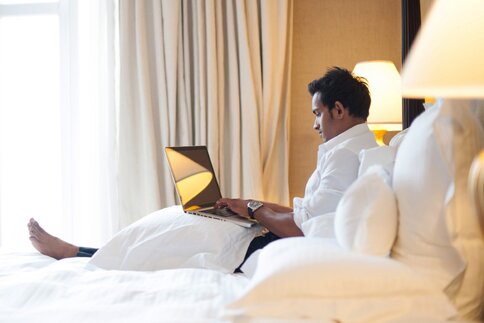 Ein Mann sitzt mit einem Laptop auf dem Schoß in einem luxuriösen Hotelzimmerbett mit weißer Bettwäsche, Schirmlampen und langen Vorhängen an den Fenstern.