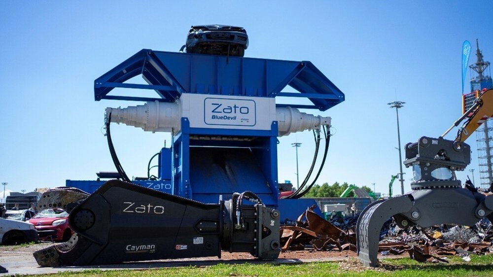 A group of people stands behind a barrier watching a live demonstration outdoors. In the foreground, the hood of a red car is open, with technical equipment next to it. Banners, parasols, and exhibition stands are visible in the background.