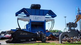 A group of people stands behind a barrier watching a live demonstration outdoors. In the foreground, the hood of a red car is open, with technical equipment next to it. Banners, parasols, and exhibition stands are visible in the background.
