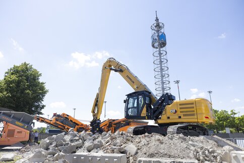 A yellow digger and an orange construction machine are standing on a mound of spoil. Behind them, the Messeturm towers into the sky.