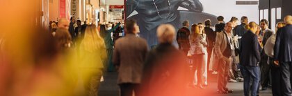 Numerous visitors to the INHORGENTA trade fair stand and walk through an exhibition room in which there is a large black and white advertising poster featuring a woman.