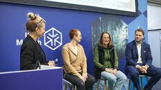 Three people are sitting at a panel discussion at ISPO Munich. A moderator stands next to them at a lectern.