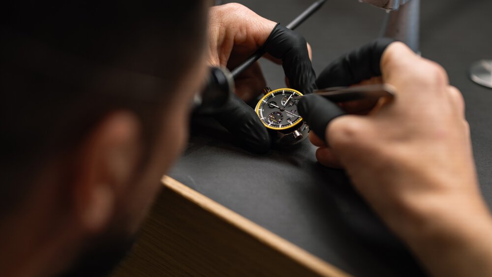  A man with black rubber gloves on his hands holds a black watch and adjusts the hour hand with tweezers.