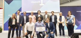 Eine Gruppe von Menschen posiert auf der Bühne der munich_i. Ein Mann in der Mitte hält einen großen Gewinnscheck in die Kamera.