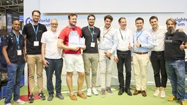 Eine Gruppe von Männern posiert mit Urkunden bei einer Siegerehrung auf der World of Quantum 2025 in München und zeigt Daumen hoch.