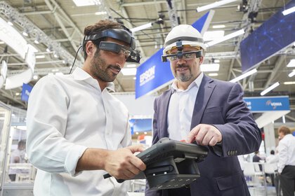 Two men wearing augmented reality goggles and hard hats use an industrial-looking tablet in an exhibition hall.