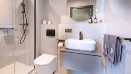 Modernes Badezimmer mit Glasdusche, wandmontierter Toilette, Waschbecken, Spiegel und grauen Handtüchern auf einem Ständer.