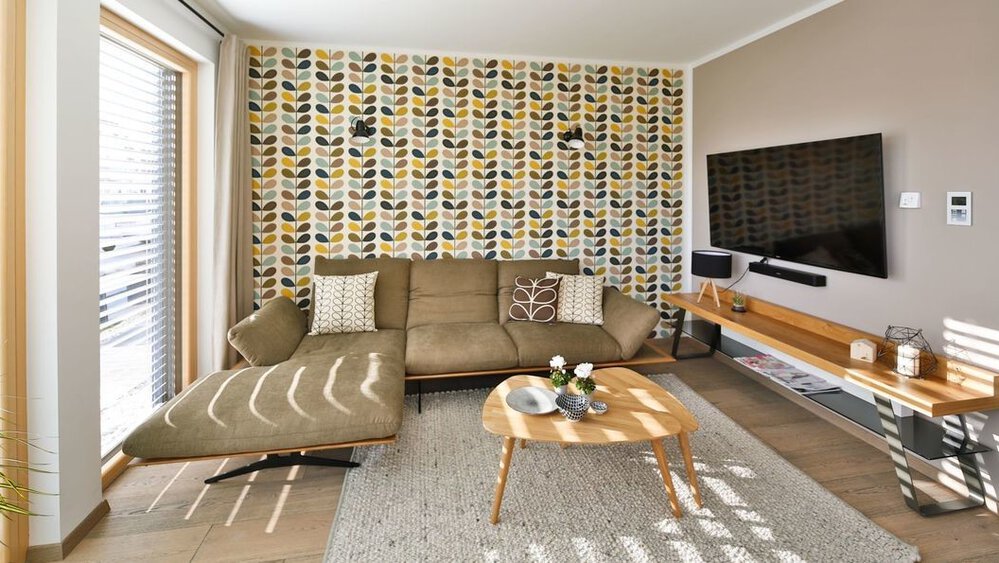 Ein modernes Wohnzimmer mit gemusterter Tapete, Sofa, Fernseher auf einem Ständer und einem runden Couchtisch auf einem gestreiften Teppich.