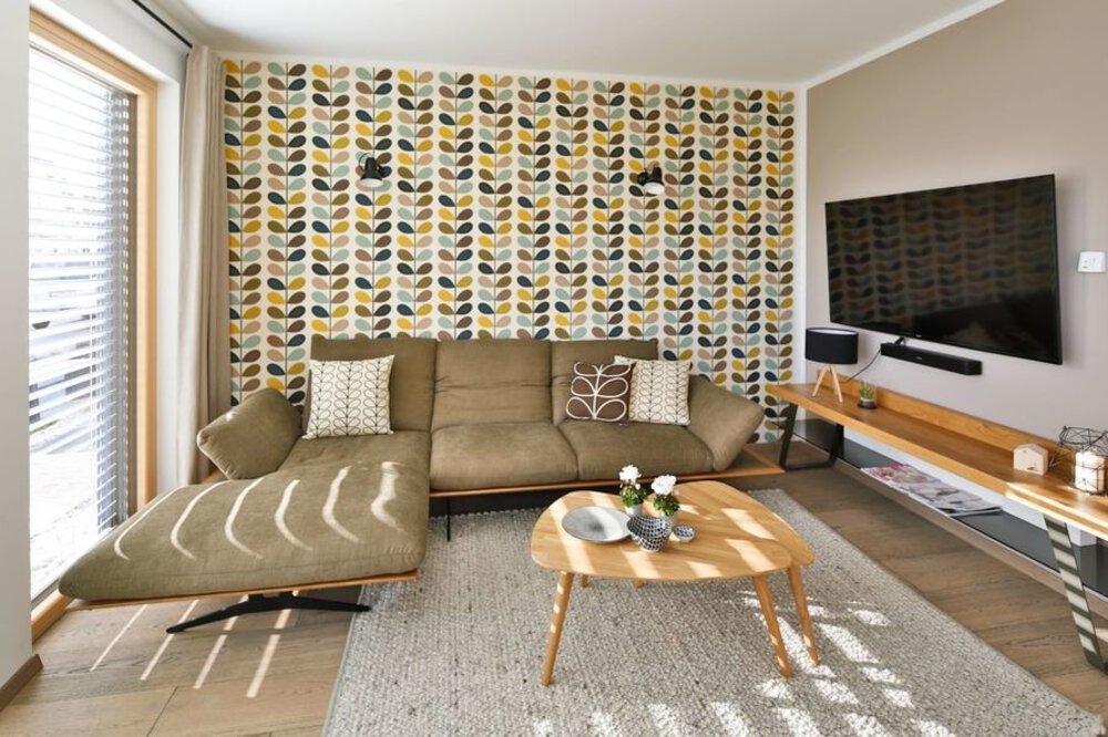Ein modernes Wohnzimmer mit gemusterter Tapete, Sofa, Fernseher auf einem Ständer und einem runden Couchtisch auf einem gestreiften Teppich.
