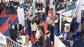 A busy exhibition hall at the EXPO REAL Career Day, with many attendees engaging in conversations at various company booths. People are gathered around standing tables, networking, and learning about opportunities. The booths are modern in design, and red and blue carpeted walkways organize the space.