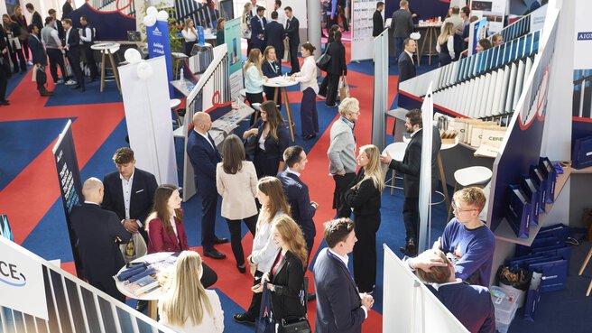 A busy exhibition hall at the EXPO REAL Career Day, with many attendees engaging in conversations at various company booths. People are gathered around standing tables, networking, and learning about opportunities. The booths are modern in design, and red and blue carpeted walkways organize the space.