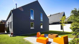 Modernes dunkelgraues Haus mit hohen Fenstern, Sitzgelegenheiten im Freien mit orangefarbenen Stühlen und einer Feuerstelle an einem sonnigen Tag.