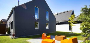 Modernes dunkelgraues Haus mit hohen Fenstern, Sitzgelegenheiten im Freien mit orangefarbenen Stühlen und einer Feuerstelle an einem sonnigen Tag.