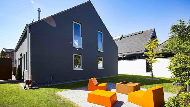 Modernes dunkelgraues Haus mit hohen Fenstern, Sitzgelegenheiten im Freien mit orangefarbenen Stühlen und einer Feuerstelle an einem sonnigen Tag
