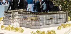 Architekturmodell eines modernen Bürogebäudes, ausgestellt an einem Messestand. Im Hintergrund sind weitere Stände und Besucher zu sehen.