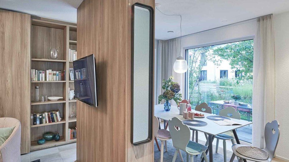 Moderner Wohnbereich mit Holztrennwand, montiertem Fernseher, Bücherregal und Esstisch neben einem großen Fenster mit Blick ins Grüne.