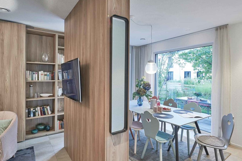 Moderner Wohnbereich mit Holztrennwand, montiertem Fernseher, Bücherregal und Esstisch neben einem großen Fenster mit Blick ins Grüne.