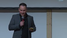 Eine Person im grauen Anzug hält ein Mikrofon und spricht vor einer Leinwand.