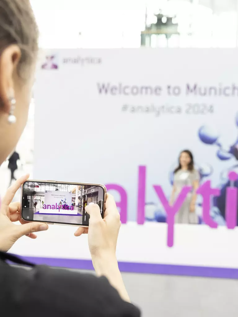 A person is holding a smartphone taking a photo at a trade fair. The word 'analytica' can be seen in purple letters. The background is blurred and you can see a banner with the text 