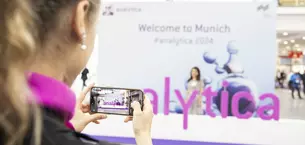 Eine Person hält ein Smartphone, das ein Foto auf einer Messe aufnimmt. Das Wort ‘analytica’ in lila Schrift in großen aufgestellten Buchstaben ist zu sehen. Der Hintergrund ist unscharf und man sieht ein Banner mit dem Text “‘Welcome to Munich #analytica 2024“.