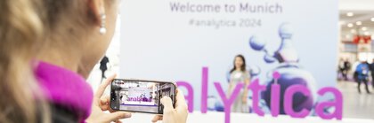 A person is holding a smartphone taking a photo at a trade fair. The word 'analytica' can be seen in purple letters. The background is blurred and you can see a banner with the text 