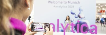 A person is holding a smartphone taking a photo at a trade fair. The word 'analytica' can be seen in purple letters. The background is blurred and you can see a banner with the text 