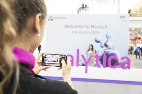 A person is holding a smartphone taking a photo at a trade fair. The word 'analytica' can be seen in purple letters. The background is blurred and you can see a banner with the text 