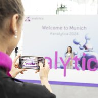 A person is holding a smartphone taking a photo at a trade fair. The word 'analytica' can be seen in purple letters. The background is blurred and you can see a banner with the text 