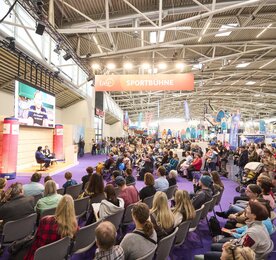 Large audience during an interview on the “Sportbühne” at the f.re.e trade fair, with a guest on stage and sports fair stands in the background.