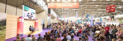 Großes Publikum bei einem Interview auf der „Sportbühne“ der Messe f.re.e, mit einem Gast auf der Bühne und Sportmesse-Ständen im Hintergrund.