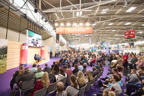 Großes Publikum bei einem Interview auf der „Sportbühne“ der Messe f.re.e, mit einem Gast auf der Bühne und Sportmesse-Ständen im Hintergrund.