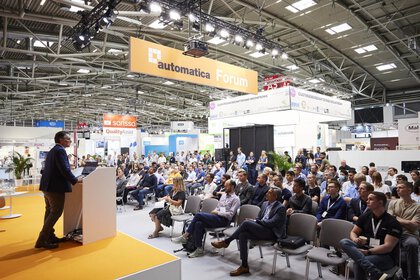 A speaker addresses a large audience from a lectern on the automatica Forum stage.