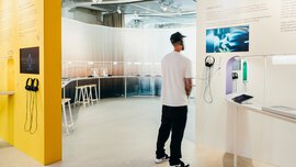 A man wearing headphones stands in front of an information wall and watches a film. In the background, various stages of watchmaking are set up.