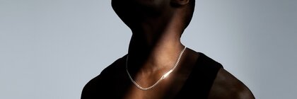 A man in a black tank top stands in the shade, tilting his head back to show off a brightly lit silver necklace.