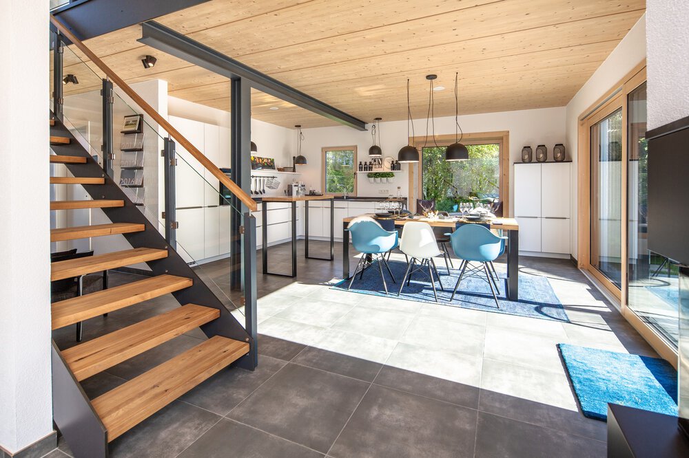 Offene Küche mit Essbereich, Holztreppe, moderner Einrichtung und großen Fenstern, die Tageslicht hereinlassen.