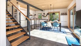 Offene Küche mit Essbereich, Holztreppe, moderner Einrichtung und großen Fenstern, die Tageslicht hereinlassen.