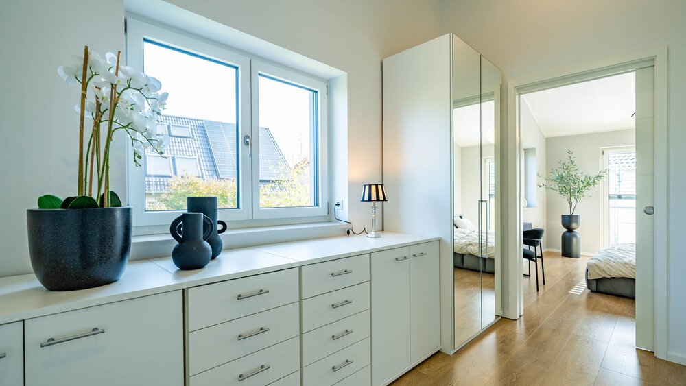 Heller Flur mit weißen Schränken, einer Orchidee im Topf, dekorativen Vasen, Spiegeltüren und einem Blick in ein Schlafzimmer mit moderner Einrichtung und Holzböden.