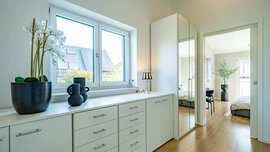 Heller Flur mit weißen Schränken, einer Orchidee im Topf, dekorativen Vasen, Spiegeltüren und einem Blick in ein Schlafzimmer mit moderner Einrichtung und Holzböden.