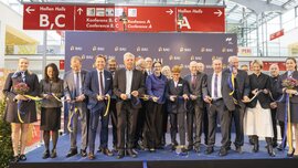 Eine Gruppe von gutgelaunten Personen in festlicher Kleidung schneidet das Eröffnungsband zur BAU Messe durch und posiert für ein Foto.