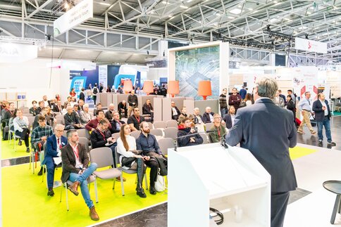 Blick von einer Bühne der electronica ins Publikum. Ein Sprecher lehnt am Podium und hält einen Vortrag. Im Hintergrund sind Messebesucher zu sehen.