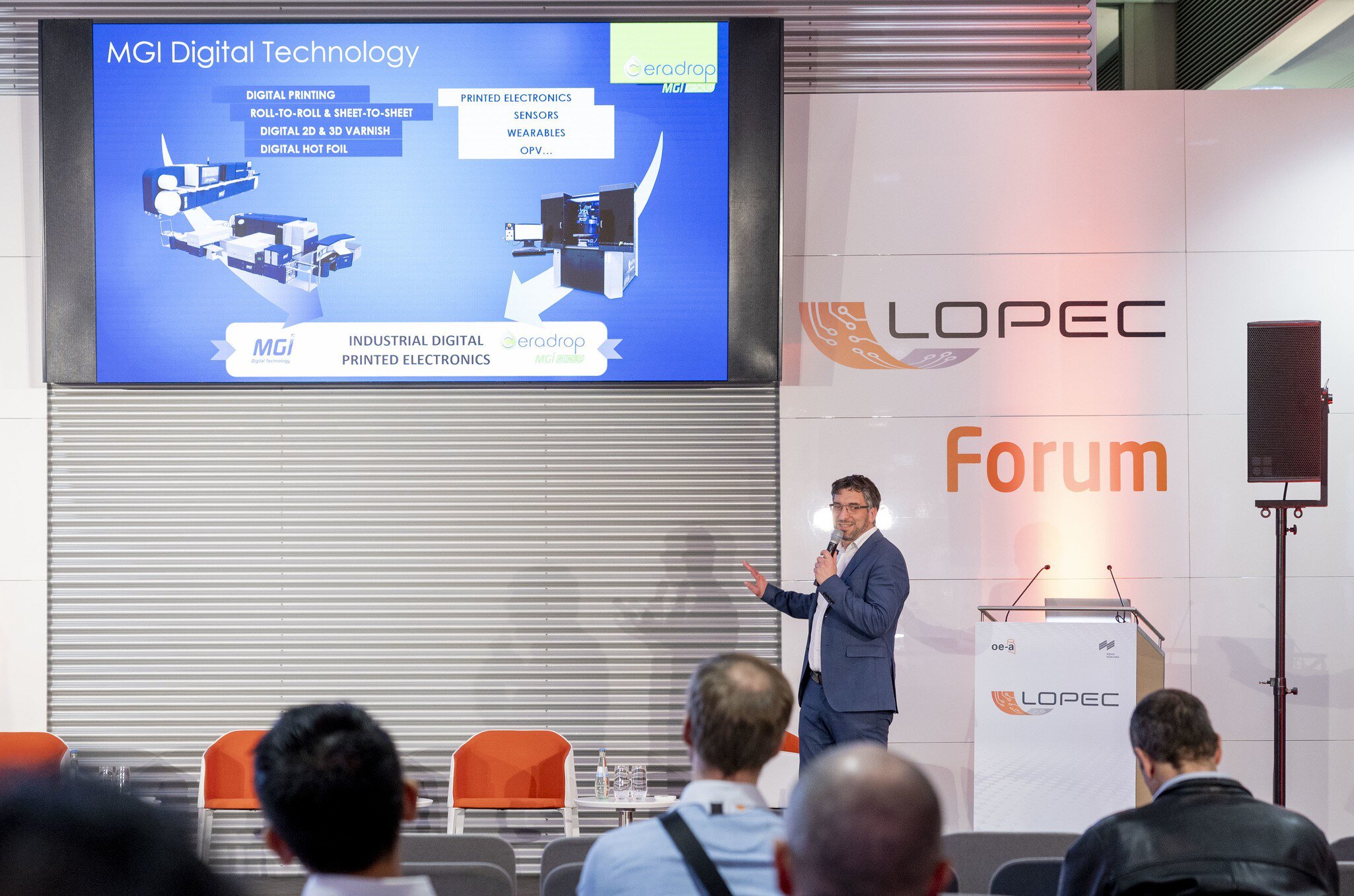 Ein Mann hält beim LOPEC Forum eine Präsentation auf einer Bühne, während ihm ein interessiertes Publikum zuhört.