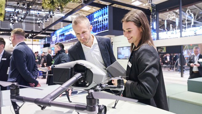 Two people look at a drone at the BAU 2025 trade fair. Other trade fair visitors can be seen in the background.