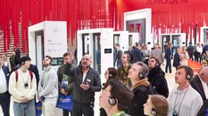 Eine Gruppe Messebesucher lauscht über Kopfhörer einem Mann mit Ansteckmikro. Sie stehen in einer Halle der BAU, in der im Hintergrund rote Bänder von der Decke hängen.