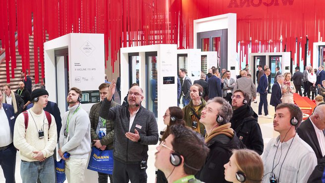 A group of trade fair visitors take part in a guided tour with headphones. In the background, exhibits can be seen between curtains of red strips hanging from the ceiling.