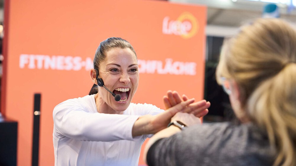 Eine Frau mit Headset und weißem Hemd steht einer anderen Frau gegenüber und hat den Mund weit aufgerissen. Ihre zusammengelegten Hände berühren einander.